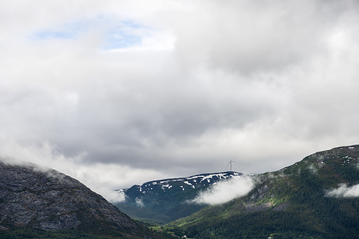 Overlooking a verdant valley, a single wind turbine stands atop a mist-covered mountain range under the vast, cloud-streaked skies of Northern Europe