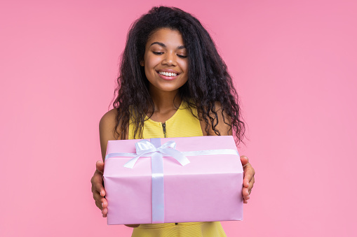 Studio shot of smiling beautiful birthday girl looking at the decorated present box in her hands with content face expression, isolatd over pastel pink background. Focus is on the gift box.