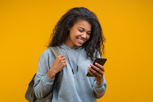 Half-length studio portrait of beautiful casually dressed girl student with perfect charming smile texting message via her mobile phone isolated on bright colored yellow background.