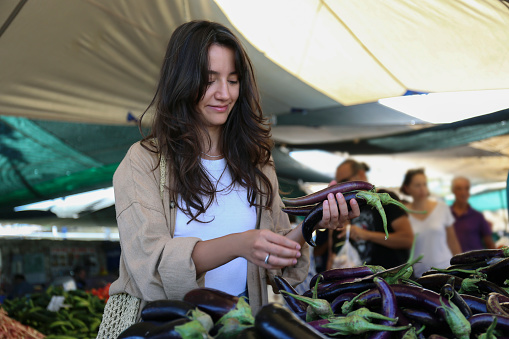 Young woman with a net bag picking out fresh eggplants on farmers market. Shopping for organic local produce vegetables. Close up, copy space, background.