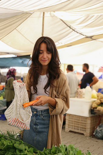 Young woman with a net bag buying fresh danvers carrots on farmers market. Shopping for organic local produce vegetables. Close up, copy space, background.