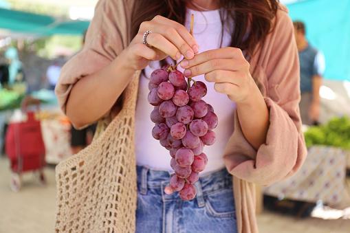 Cropped shot of a young woman with a net bag picking fresh grapes on farmers market. Shopping for organic local produce fruits. Close up, copy space, background.