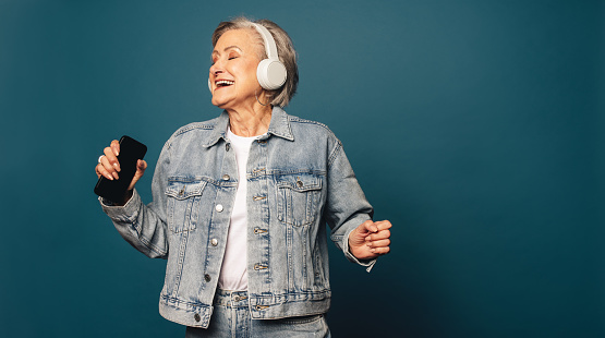 Happy, fashionable senior citizen in casual denim clothing stands in a studio, holding a smartphone and wearing headphones. She listens to music, showcasing her love for entertainment and technology.