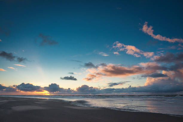 A sunset on the beach from the North Sea island of Juist in spring with almost clear skies. Slowly the waves roll on the wide beach of Juist. In the sky, only a few clouds can be seen on the horizon. The setting sun shines through between them. More clouds shine in the light of the setting sun.