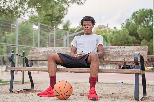 young basketball player waiting sitting on a bench with a ball between his legs