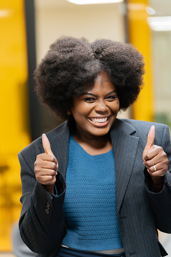 Smiling businesswoman with afro hair gesturing thumbs up in a corporate setting, exuding positivity.