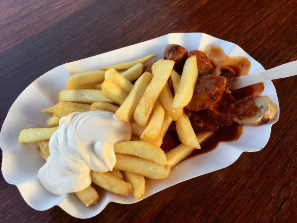 Close-up of a traditional German currywurst served with spicy curry sauce and bread on a white paper tray, photographed outdoors in sunlight.