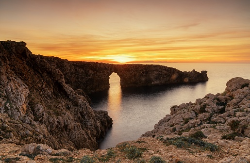 A view of the landmark stone arch of Pont d'en Gil on Menorca Island at sunset