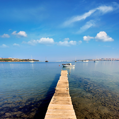 Image of a wooden brown pier with ropes near amazing blue sea water near rocks on the background of many boats on a cloudless fall day. Cyprus, Konnos beach