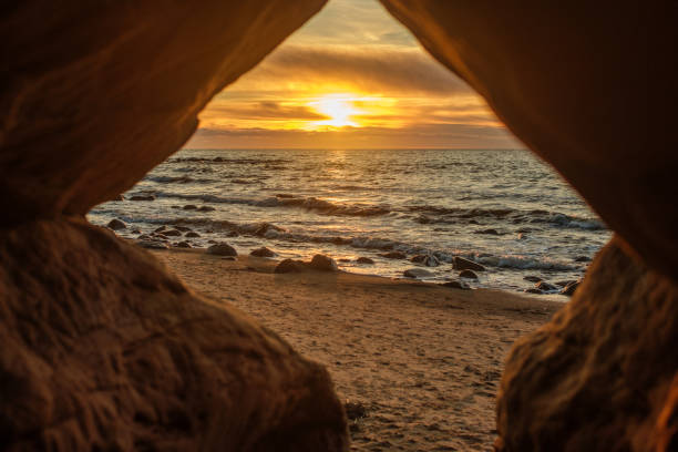 A magical moment unfolds as the sun dips below the horizon, illuminating the cave at Veczemju Klintis Beach with its golden light, creating a mesmerizing silhouette against the evening sky.
