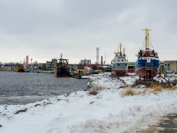 Braving the chill of a cold winter day, fishing boats rest along the coast of Ventspils, their colorful hulls contrasting against the snowy backdrop, a testament to the enduring spirit of maritime life in the Baltic.