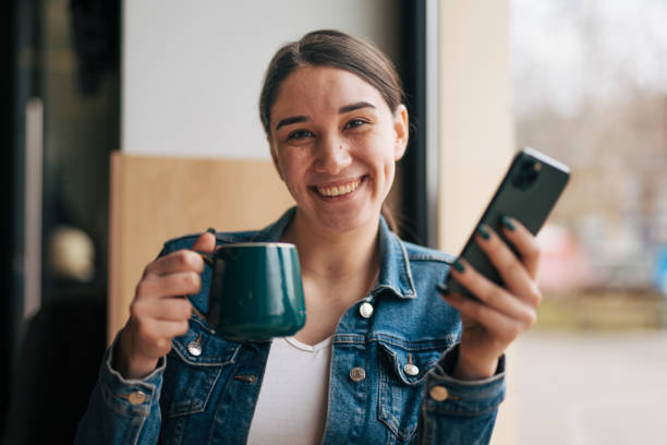 Young smiling girl holding a cup of hot drink and using her smart phone, enjoying in coffee bar.