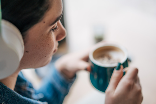 Cheerful young woman drinking coffee and wearing wireless headphones while listening to music in a cafe.Positive beautiful girl sitting and relaxing in coffee shop.