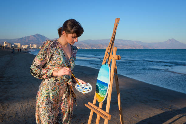 A woman is standing on a sandy beach next to an easel, with the ocean in the background. She appears focused on painting a scene from the beach.