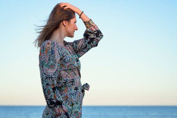 A woman with long hair is standing on the sandy beach, facing the ocean. She is wearing a bathing suit and looks out at the water under the clear sky.
