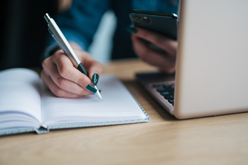 Close-up photo of freelance girl writes in a notebook and uses a laptop and phone in a cafe