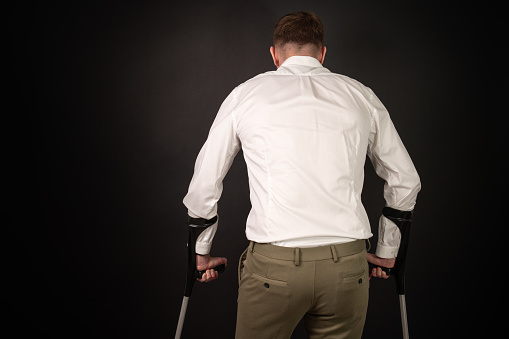 Man in white shirt looking away being sad while using crutches, seen from behind.