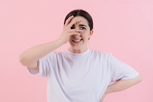 Scared attractive Caucasian young brunette woman in casual clothes covered her face with her hand and looking at the camera isolated on a pink studio background