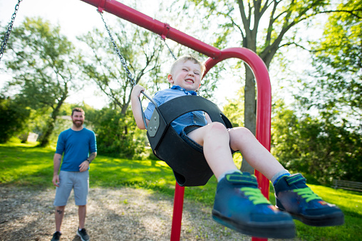 Little boy has his eyes closed as he is laughing and smiling with excitement as his dad pushes him high on the swing set at the playground outdoors in the summer.