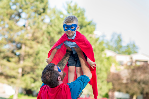Cute little boy in a blue superhero mask and red cape is matching his father as they play pretend superheroes at the park outdoors in the summer. The young boy is laughing and looking at the camera as his father is holding him up to the sky so he can pretend he is flying to build creativity and have fun.