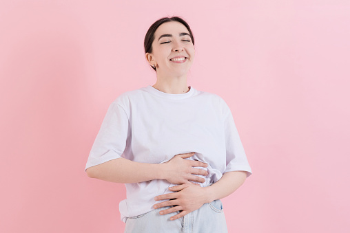 Caucasian young brunette woman in casual white t-shirt smiling and touching her stomach after a delicious healthy eating closing her eyes isolated on pink studio background