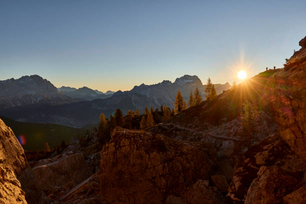 Mountain and Larches trees on a sunny day in the Dolomites, the sky is clear and the sun is shining just above the horizon of the mountains