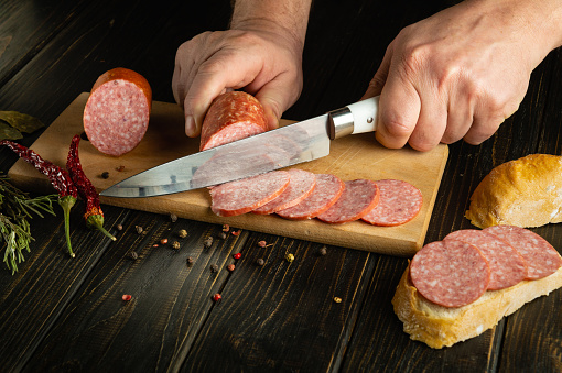 The process of preparing sandwiches with bread and sausage on the kitchen table for a snack. Slicing meat sausage with a knife in the hand of a cook.