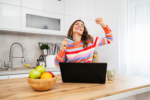 Beautiful and happy housewife preparing for new meal cooking. She is using her laptop and credit card for online food ingredient shopping or purchasing.