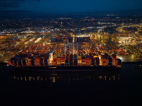 Side view of container ship with lights at night. High altitude aerial view of illuminated Southampton Docks and urban Southampton city. Lights reflection on the sea.