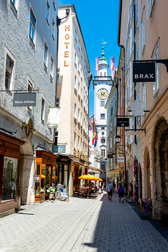 Salzburg, Salzburg - Austria - 06-18-2021: Bustling Salzburg alley with tourists, shops, and the iconic Old Town clock tower of city hall under a clear blue sky