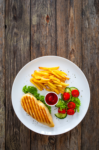 Grilled chicken breast with fries and fresh vegetables on wooden background
