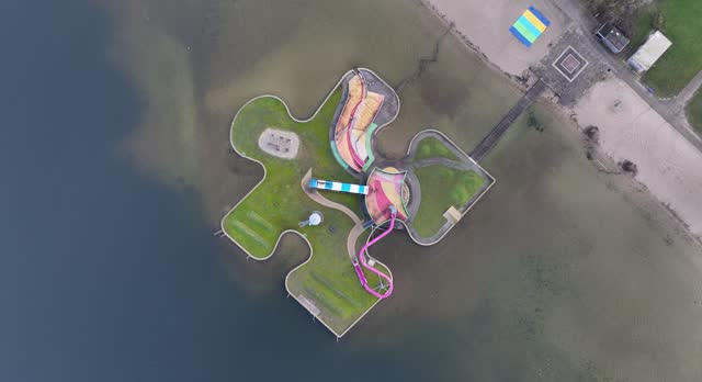 The water play island in the shape of a puzzle piece provides lots of water fun for young and old. Recreational playground swimming water surround it. Birds eye aerial drone view.