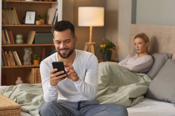 man with smartphone in bedroom, upset woman in the background - infidelity dishonesty heterosexual couple couple imagens e fotografias de stock