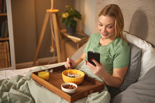 Happy young woman using mobile phone while having breakfast in bed