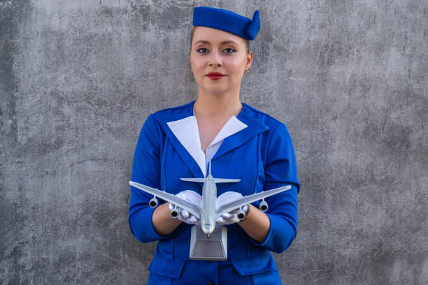 woman stewardess in blue vintage uniform with hat, gloves poses with aircraft model in hands. Flight attendant in golden age of air travel. Work in aviation at airline. Customer service industry, travel by aircraft around the world
