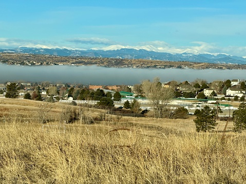 A low layer of fog settles in over the Denver suburb of Parker, Colorado.  Neighborhoods, a school, an equestrian facility and sport fields are in the foreground.  The rolling hills of the front range and snow-capped mountains of the Rockies can be seen in the background. The layer of fog appears to divide the scene between near and far.