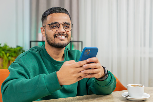 Indian man sits at table uses mobile phone smiles at home office room apartment. Smiling Arabian guy texting share messages content on smartphone social media applications online watching relax movie