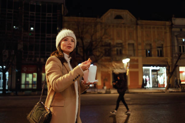 Amidst the evening's festive lights, a woman enjoys the simple pleasure of a street food meal, a delightful pause in her journey home. The steam rises from the container, mingling with the crisp night air, as she savors the taste of her quick, delicious choice — a perfect embodiment of urban life.