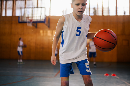 Dynamic junior basketball player in action dribbling a ball, learning and practicing moves at indoor court. A young athlete is practicing moves with basketball. Portrait of active boy with basketball.