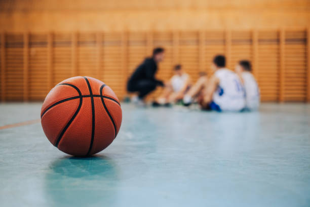 A basketball on court with junior team in blurry background. A basketball on court with a junior team with coach in a blurry background. A ball on basketball court during the training. A junior team with basketball coach in a blurry background. Copy space. youth basketball indoor stock pictures, royalty-free photos & images
