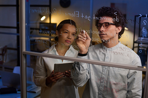 Young man and woman working together on code writing it on glass wall in IT company office late in evening