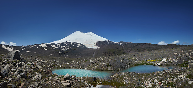 Panoramic view of a snow-capped mountain Elbrus with a turquoise glacial lake pool in the rocky foreground under a clear blue sky