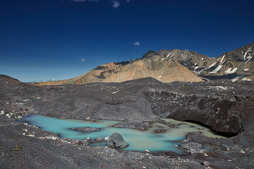 Turquoise glacial lake pool nestles among dark moraines under a stark mountain range and blue sky