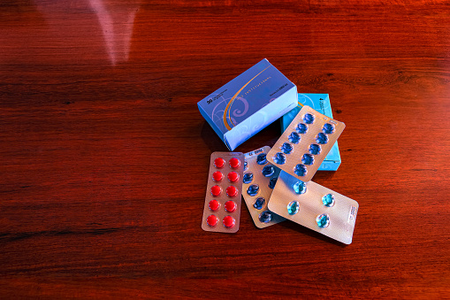 Some vitamins in blister packs on a rich shiny rosewood table. There are both Vitamin D3 in Blue and B Complex in Red. Shot in a studio environment with natural light. Long Exposure. Copy space. Note to Inspector: All logos, brand names and batch numbers have been removed.