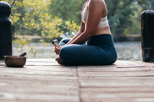Close up of an unrecognizable woman in sportswear meditating and doing yoga in nature. She burns a wooden palo santo.