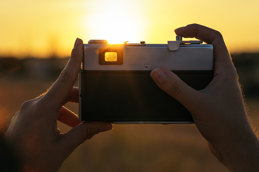 Close-up of an analog camera. An unrecognizable person holds her and takes photos in the field at sunset. Concept of analog, retro, classic and vintage.