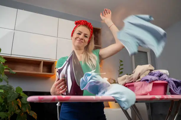 Beautiful young blonde woman with cute bow ironing at home, clothes flying around, photomanipulation