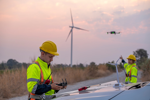 Engineer wearing uniform read blueprint document inspection work in wind turbine farms rotation to electricity,standing in front of car Model windmill and wind force meter is ecology energy concept.
