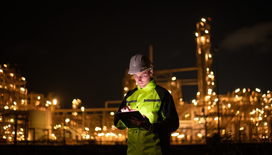 Engineer survey team wear uniform and helmet stand workplace checking blueprint project and radio communication inspection work construction site with night lights oil refinery background.