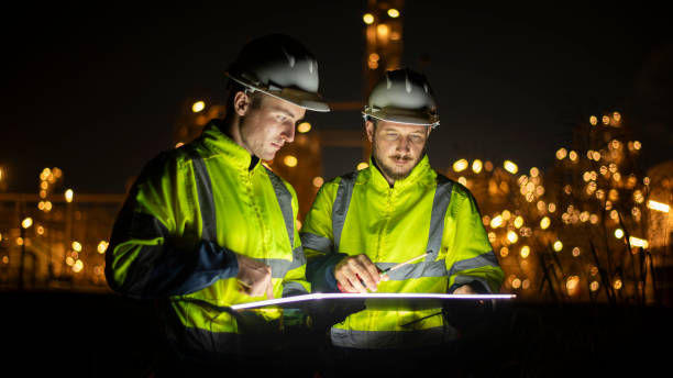 istock Engineer survey team wear uniform and helmet stand workplace checking blueprint project and radio communication inspection work construction site with night lights oil refinery background 2047190565